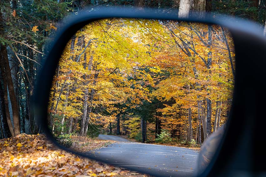 View in a car mirror of the receding highway lined with gold and orange trees.