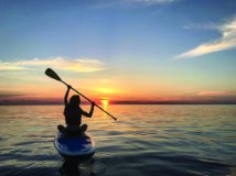 A person sitting on a paddle board with the paddle raised and the sun setting in the background