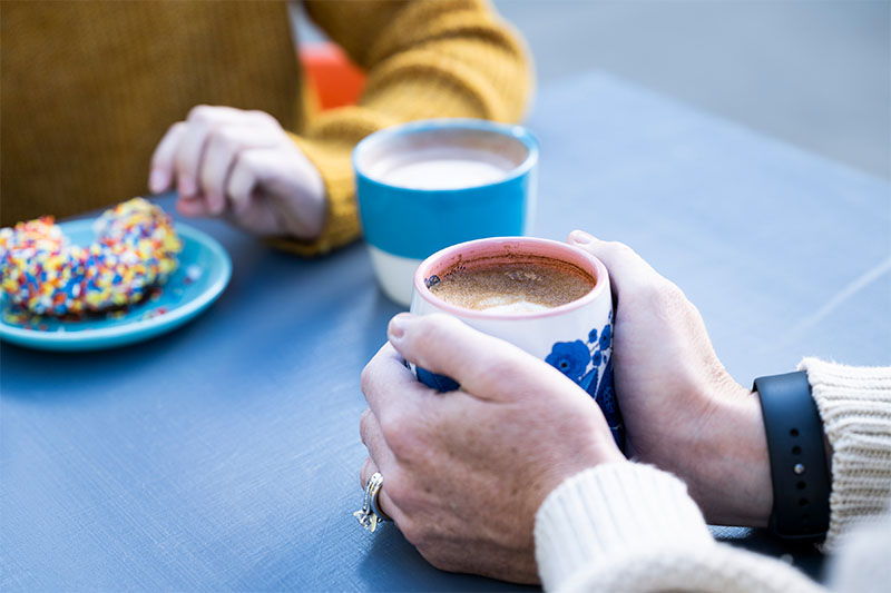 A woman cups a cup of coffee in her hands when across from her a person's hands sit beside a plate with a donut covered in sprinkles.
