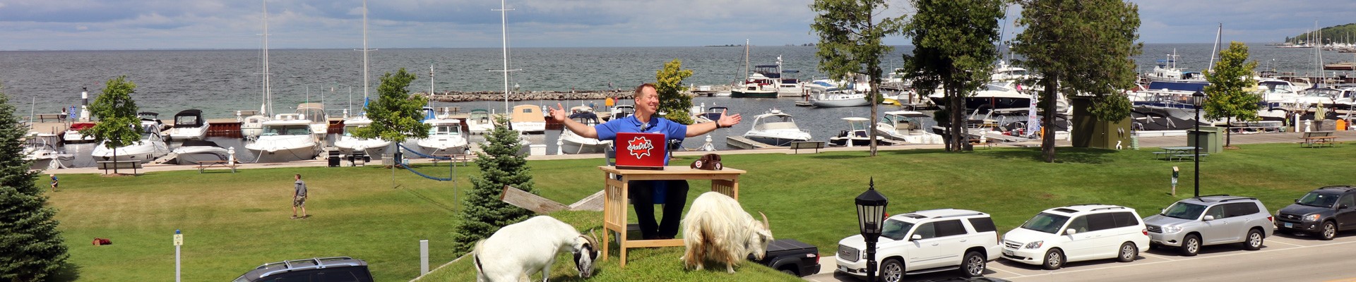 Aman with a laptop and two goats sitting at the lakefront in front of the marina