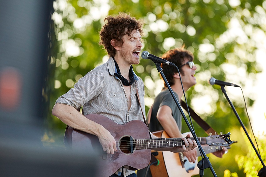 A rock band plays guitars and sings on an outdoor stage among the trees